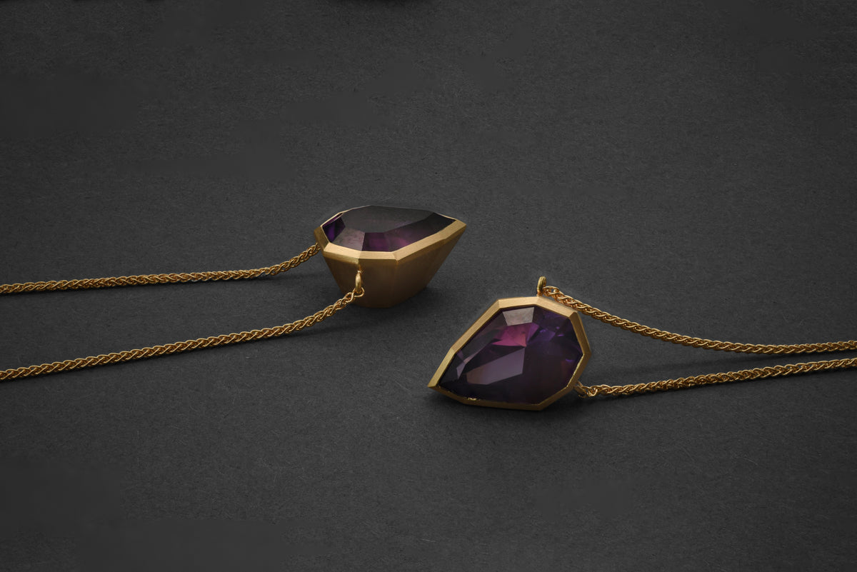 Two gold pendant necklaces with faceted dark purple amethyst, displayed on a dark gray textured background.
